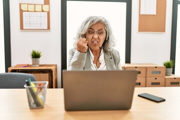 Middle age businesswoman sitting on desk working using laptop at office angry and mad raising fist frustrated and furious while shouting with anger. rage and aggressive concept.
