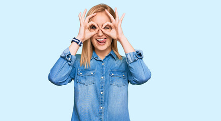 Beautiful caucasian woman wearing casual denim jacket doing ok gesture like binoculars sticking tongue out, eyes looking through fingers. crazy expression.