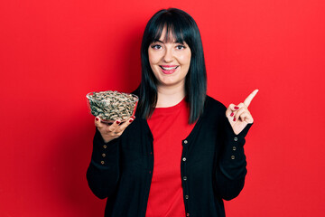 Young hispanic woman holding sunflower seeds bowl smiling happy pointing with hand and finger to the side