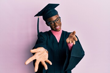 Young african american girl wearing graduation cap and ceremony robe smiling cheerful offering...