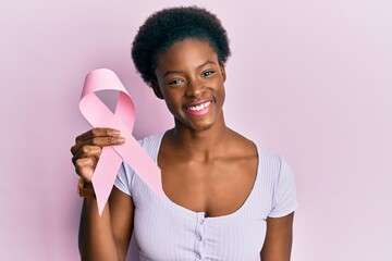 Young african american girl holding pink cancer ribbon looking positive and happy standing and...