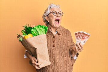 Senior grey-haired woman holding groceries and israel shekels banknotes angry and mad screaming...