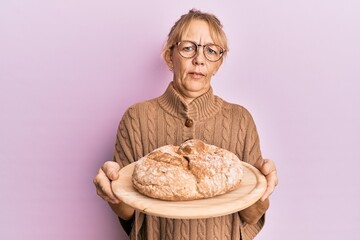Middle age blonde woman holding bread clueless and confused expression. doubt concept.