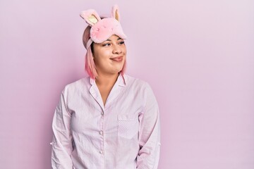 Hispanic woman with pink hair wearing sleep mask and pajama smiling looking to the side and staring away thinking.