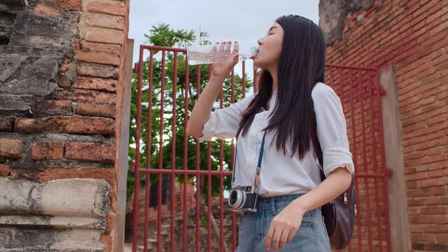 Young Asian backpacker woman blogger tourist with camera feel tired stand in front of pagoda take a rest and drink water in plastic bottle at old town , Lifestyle tourist travel holiday concept.