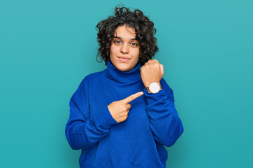 Young hispanic woman with curly hair wearing turtleneck sweater in hurry pointing to watch time,...