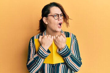 Young caucasian woman holding book angry and mad screaming frustrated and furious, shouting with anger. rage and aggressive concept.