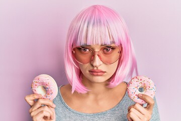 Young caucasian woman wearing pink wig holding donuts skeptic and nervous, frowning upset because...