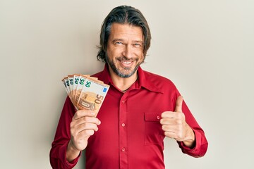 Middle age handsome man holding bunch of 50 euro banknotes smiling happy and positive, thumb up...