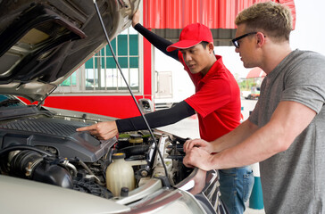 An Asian gas station worker raises the car bonnet to check for the cause of an engine malfunction. A caucasian driver with sunglass stand listening attentively in front of broken pickup truck. © chadchai