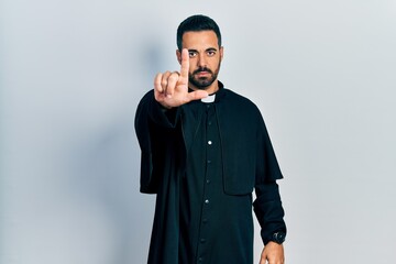 Handsome hispanic man with beard wearing catholic priest robe pointing with finger up and angry expression, showing no gesture