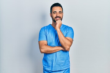 Handsome hispanic man with beard wearing blue male nurse uniform looking confident at the camera...