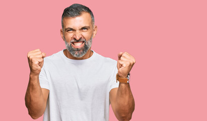 Middle age handsome man wearing casual white tshirt angry and mad raising fists frustrated and...