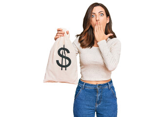 Young brunette woman holding dollars bag covering mouth with hand, shocked and afraid for mistake....