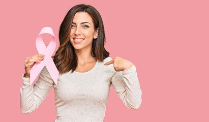 Young brunette woman holding pink cancer ribbon pointing finger to one self smiling happy and proud