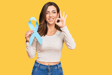 Young brunette woman holding blue ribbon doing ok sign with fingers, smiling friendly gesturing...