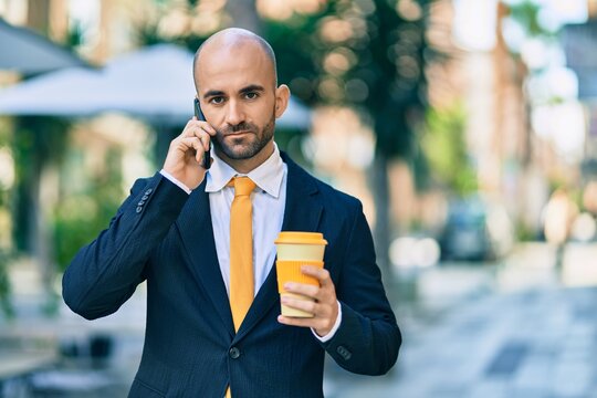 Young hispanic bald businessman with serious expression talking on the smartphone drinking coffee at the city