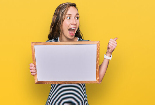Young blonde woman holding empty white board pointing thumb up to the side smiling happy with open mouth