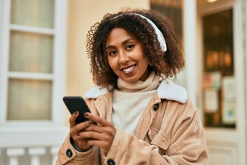 Young african american woman smiling happy using smartphone and headphones at the city.