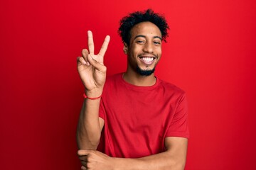 Young african american man with beard wearing casual red t shirt smiling with happy face winking at...