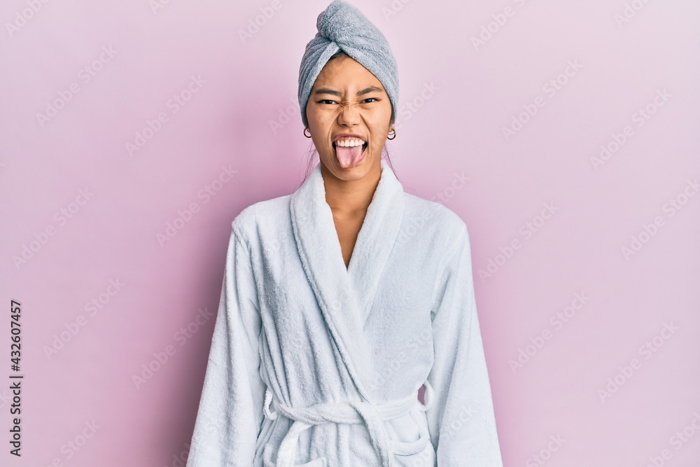 Sticker Young chinese woman wearing shower towel cap and bathrobe sticking tongue out happy with funny expression. emotion concept.