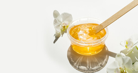 Sugar paste or wax honey in a transparent jar and white orchid on a white background. Sugaring. Depilation and beauty concept. Waxing. - obrazy, fototapety, plakaty
