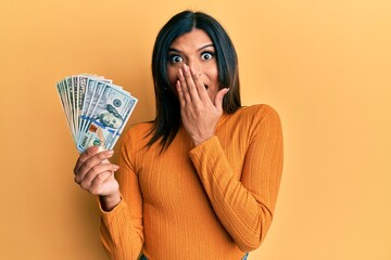 Young latin transsexual transgender woman holding dollars covering mouth with hand, shocked and...