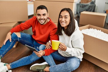 Young latin couple drinking coffee and using touchpad at new home.