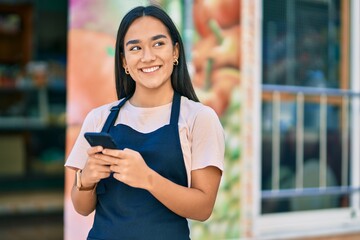 Young latin shopkeeper girl smiling happy using smartphone at fruit store.