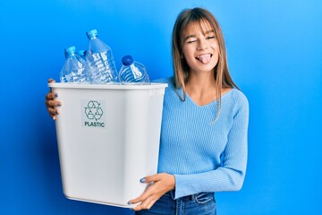 Teenager caucasian girl holding recycling bag with plastic bottles sticking tongue out happy with...