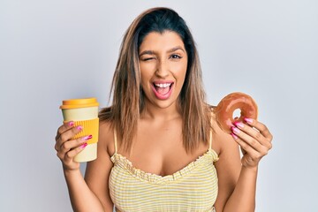 Beautiful brunette woman eating doughnut and drinking coffee winking looking at the camera with sexy expression, cheerful and happy face.