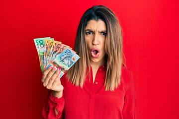 Beautiful brunette woman holding australian dollars in shock face, looking skeptical and sarcastic, surprised with open mouth