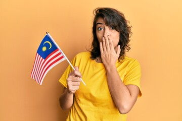 Young hispanic woman holding malaysia flag covering mouth with hand, shocked and afraid for mistake. surprised expression