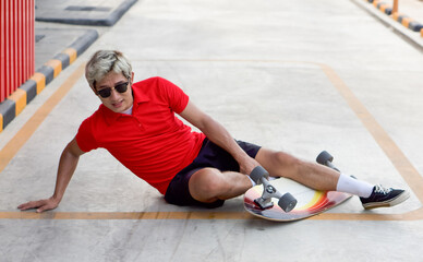 Young asian man dressed in a red T-shirt and black shorts caught his ankle due to a skateboarding accident. He lifted himself with one hand from the ground after the crash.