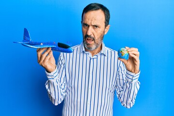 Middle age hispanic man holding plane toy and world ball in shock face, looking skeptical and...