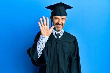 Middle age hispanic man wearing graduation cap and ceremony robe showing and pointing up with fingers number five while smiling confident and happy.