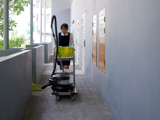 Fototapeta na wymiar Young hotel maid in blue uniform push housekeeping cart on the corridor in front of the hotel room. The laundry basket, Glass cleaner, Rubber gloves, Door mat and Vacuum cleaner are on the cart.