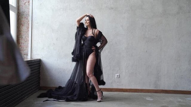 sexy woman in black bodysuit and negligee stands at gray wall. photo shoot