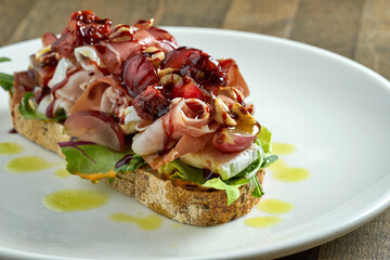 Huge sandwich with jamon crudo, brie cheese, mixed salad, grapes, sun-dried tomatoes and nuts on rye bread, served in a white plate on a wooden background. Delicious breakfast toast