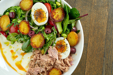 Classic nicoise salad with boiled egg, potatoes, tuna, olives and white sauce, served in a white bowl on a wooden plate. A delicious and healthy seafood salad