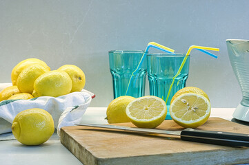 Tabla de madera con limones cortados para ser exprimidos, al fondo hay más limones, dos vasos y un...