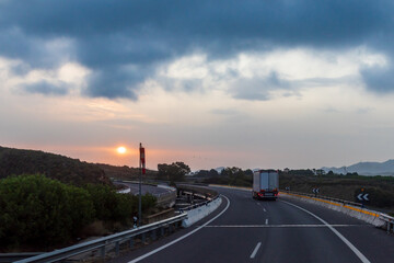 Fototapeta na wymiar Truck driving on the highway next to a windsock and the sun peeking through the clouds at dawn.