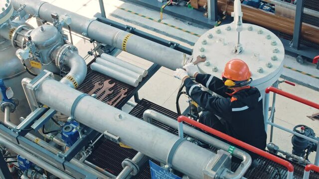 The mechanic is dressed in a black uniform, orange hard hat, goggles and earmuffs. Tightens the nuts with a large wrench on the pressure vessel. Work at an oil refinery.