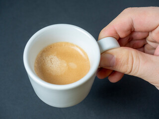 a man's hand holds a white cup of freshly brewed espresso on a dark background. Brown foam. An invigorating drink. top view