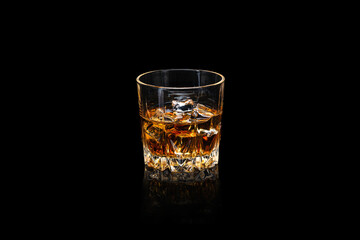 Glass of whiskey with ice cubes on a dark reflective surface isolated on black.