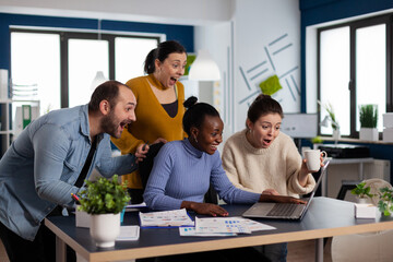 Startup diversity team happy celebrating after agreement signing with important client, partnership looking at laptop. Cheerful multiethnic business team with laptop and papers excited about project.