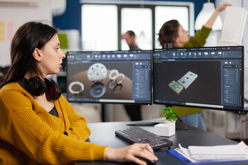 Industrial female engineer looking at personal computer with dual monitors setup, screens showing CAD software with 3D prototype of gears metalic mechanical piece. Employee working in creative office