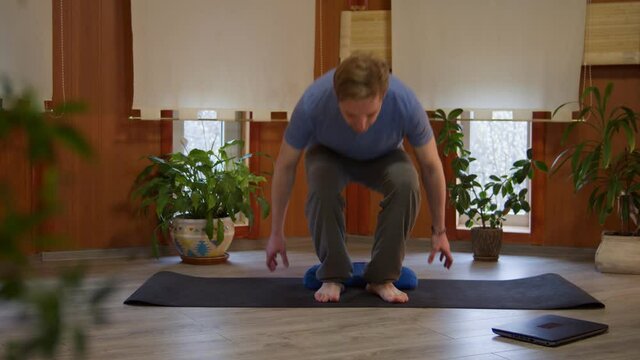 Handsome young man approaching yoga mat, switching on laptop to be ready doing yoga via internet in living room at home, nice-looking male sitting on floor using meditation cushion in bedroom, potted