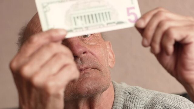 Close-up of a Caucasian pensioner examines a 5 dollar bill, holding it in front of his eyes. 5 dollar banknote in the hands of a man. selective focus, shallow depth of field
