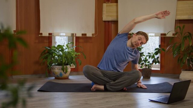 Front view of fit young man doing stretching hand exercises with online yoga trainer via internet, sporty male practicing yoga asana using laptop computer and live stream video classes, remote home